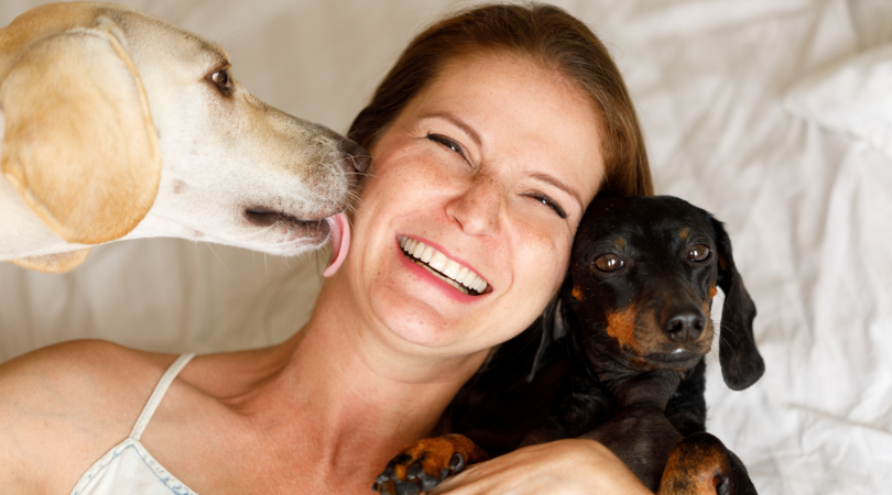 A lady getting a kiss from one dog while another dog looks at her on her bed.