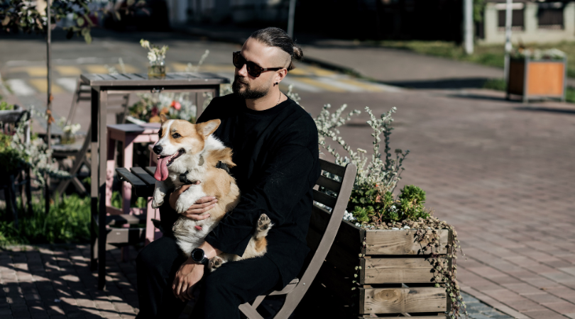 A hipster man sits at a cafe table with a small dog comfortably nestled on his lap. The man looks relaxed and content, enjoying the company of his furry friend in the laid-back atmosphere of the pet-friendly cafe.